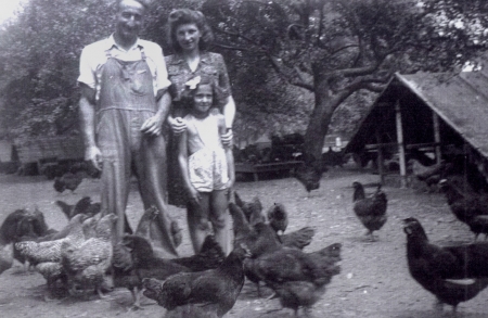 Black-and-white photograph of a man and woman with a young child with curly blond hair. They stand together outdoors in front of a truck and a barn in the background.