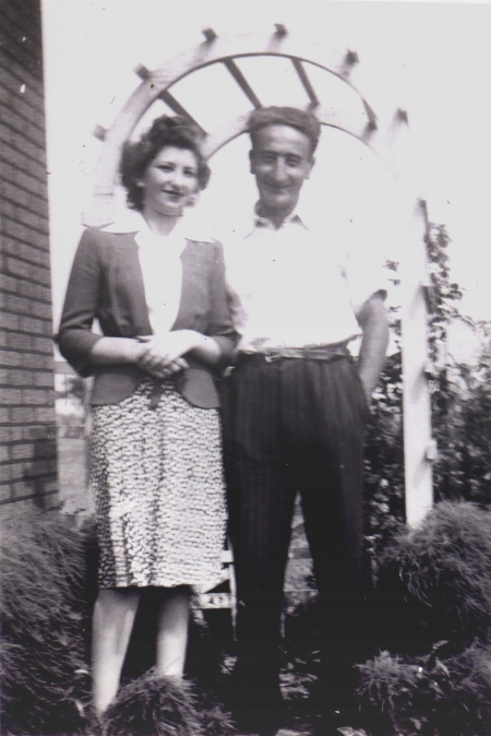 Black-and-white photograph of a man and woman standing together under an arbour outdoors, smiling at the camera. The man has his hands in his pockets.