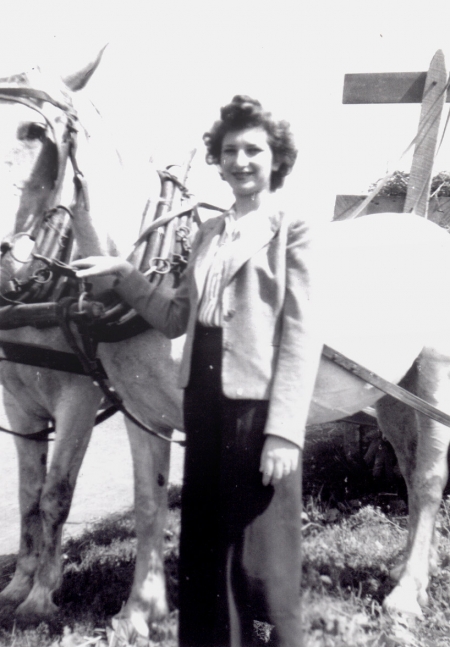 Black-and-white photograph of woman with dark brown hair, standing outdoors beside two white horses tied up to a carriage or wagon. She rests one of her hands on the horse’s reins.