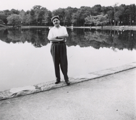 Black-and-white photograph of a man standing with his arms crossed, in front of a large pond or lake. He wears a short-sleeved shirt, dark trousers, and a cap. Trees line the lake in the background.