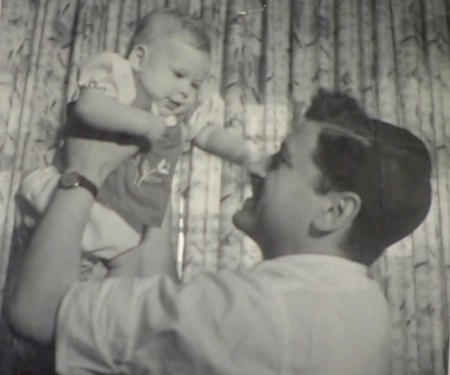 Black-and-white photograph of the side profile of a man holding up and looking at a baby in his arms. The baby looks down at him with her arms stretched out, smiling at the man.