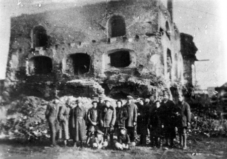 Photo en noir et blanc d'un groupe de 15 personnes se tenant ensemble en ligne à l'extérieur, deux enfants sont assis sur le sol, devant les ruines d'un bâtiment. Le bâtiment n'a plus de toit et plusieurs cadres de fenêtres sont sans fenêtres.