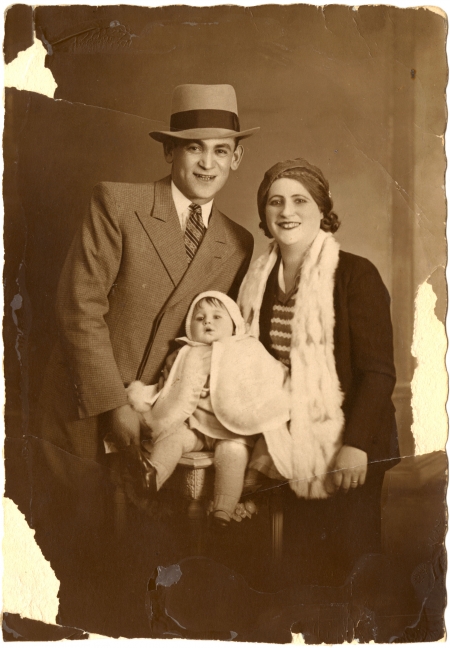 Sepia-tone photograph of a man and woman posing with a baby for a portrait. The man wears a hat and suit and tie, and the woman also wears a hat. The baby wears a white cape. The photograph’s edges have some wear and tear.