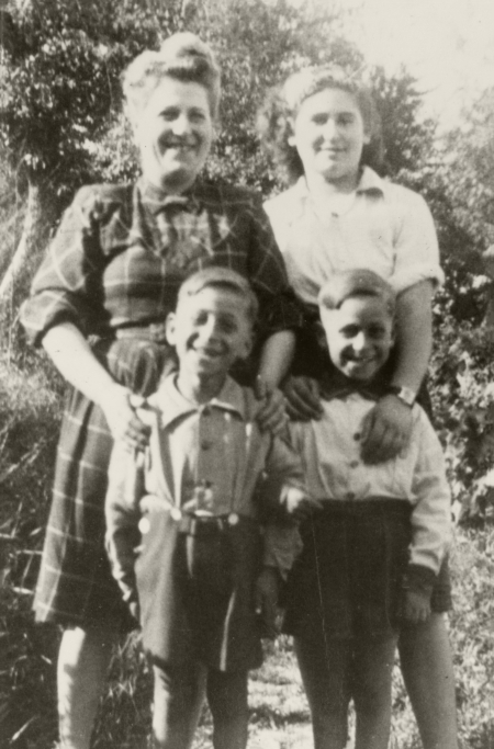 Black-and-white photograph of a two women standing behind two young twin boys, all 4 of them smiling at the camera. They are standing outside and in what looks like summer weather, due to the women’s rolled-up sleeves, and the boys’ shorts.