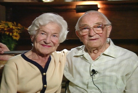 Photo en couleur d'un couple de personnes âgées assis ensemble bras-dessus bras-dessous et souriant à la caméra. La femme sur la gauche porte un cardigan et l'homme porte des lunettes et une chemise à col.