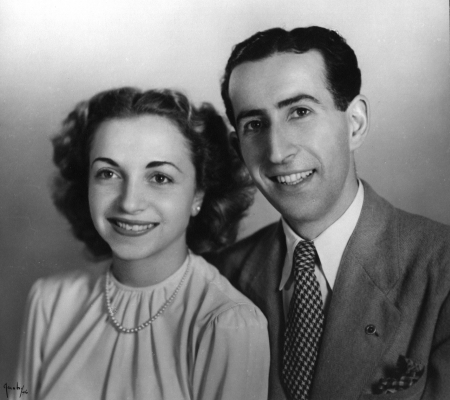 Black-and-white studio photograph of a man and woman sitting together and smiling at the camera. The man wears a suit, and the woman wears a blouse with a pearl necklace. Both have dark brown hair.