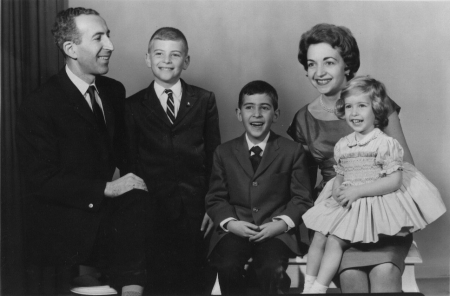 Black-and-white studio photograph of a man and woman with their three young children, smiling at the camera. The man and two boys wear suits, and the woman and her daughter are in dresses.