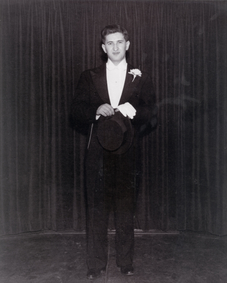 Black-and-white photograph of a young man standing in a tuxedo, in front of a dark curtain.