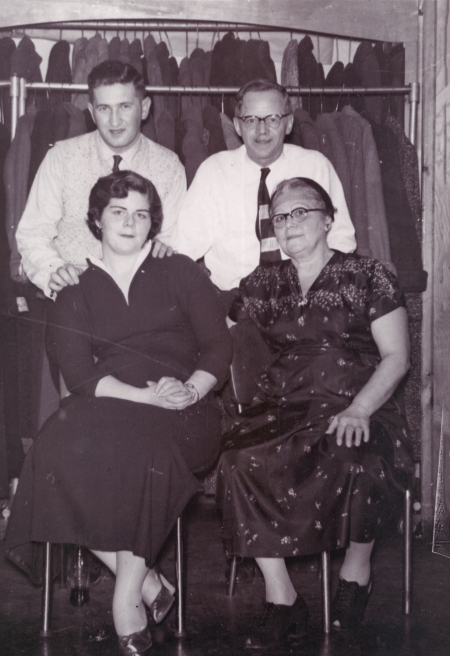 Black-and-white photograph of two men standing behind two women, who are sitting on chairs. The couple on the right appears to be older than the other couple. Both men wear shirts and ties, and the women wear dresses.