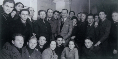 Photo en noir et blanc d'un grand groupe d'environ trente jeunes, rassemblés en deux rangées et souriant à la caméra. Un homme plus âgé portant un complet est debout au centre du groupe.