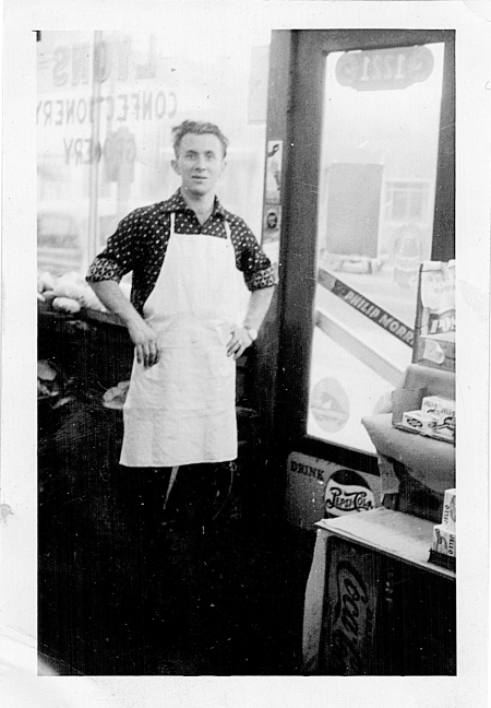 Photo en noir et blanc d'un homme se tenant à l'intérieur d'une épicerie. Il a les mains appuyées sur ses hanches et il porte un tablier blanc, il est debout près de la porte d'entrée du commerce. Il y a les mots “LYONS CONFECTIONARY STORE” écrits à l'envers dans la fenêtre derrière lui.