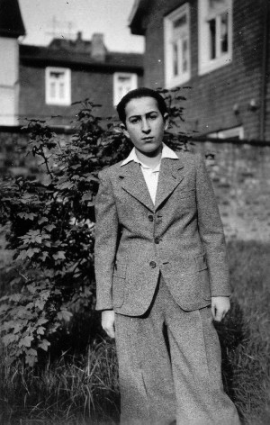 Black-and-white photograph of a young man standing outdoors in what appears to be a backyard, with a fence and shrub behind him. He wears a suit jacket and collared shirt, with his hair combed back.