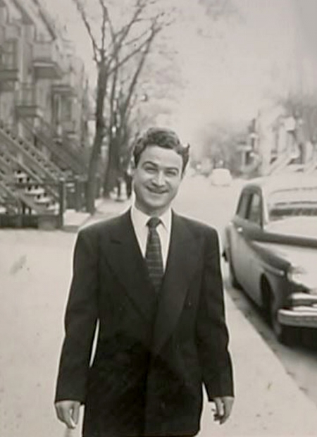 Black-and-white photograph of a man standing outside on a sidewalk, wearing a suit and smiling at the camera. The street behind him has a vintage car parked on the right, as well as trees, balconies, and stairwells leading up to apartments.