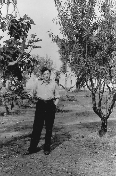 Black-and-white photograph of a young man standing on a lawn, with a tree on either side of him. He has his hands in his pockets, and wears dark trousers and a light collared shirt.