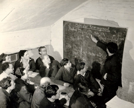 Black-and-white photograph of a group of about a dozen men looking at a chalk board. One of the men stands with a piece of chalk and points at an equation written on the board. The other men are sitting at a table, some taking notes with pen and paper.