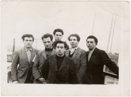 Photo en noir et blanc d'un groupe de six jeunes hommes habillés de manteaux debout ensemble sur le pont d'un navire.