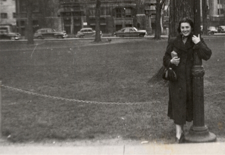 Photo en noir et blanc d'une femme souriant et s'appuyant sur un lampadaire devant un grand champ avec des bâtiments et des voitures en arrière-plan. Elle tient un sac-à-main et un livre dans sa main.