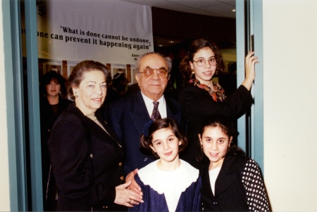 Photo en couleur d'un homme et d'une femme âgés se tenant ensemble à l'intérieur avec une autre femme et deux jeunes filles. La famille est habillée de manière formelle et les filles portent des blouses et l'homme un complet.