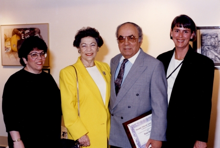 Colour photograph of an elderly man and woman standing between two other women. The group of four smile at the camera. The man wears a suit and holds a framed document.