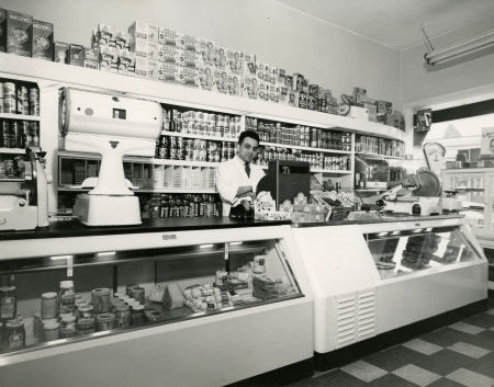 Black-and-white photograph of a man standing behind the cash register of  a large grocery store. He stands in front of a shelf full of food products, and there is a large display food counter in front of him.