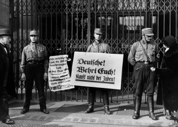 Black-and-white photograph of three men in military uniforms, standing in front of a closed storefront that has iron gates. Two of the hold up signs written in German. The other soldier is in discussion with a civilian.