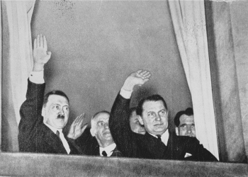 Photo en noir et blanc de deux hommes saluant à partir d'une fenêtre. La photo est prise d'en bas de l'extérieur. L'homme à gauche porte une moustache, et il y a des hommes derrière eux.