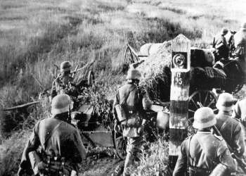 Black-and-white photograph of a military troop of about half a dozen men walking into a field. There appears to be a large fire with a cloud of dark smoke in the far distance.