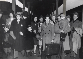 Black-and-white photograph a group of about 16 people walking towards the camera on a train platform. They are wearing jackets and some carrying suitcases. There are four young children in the group.