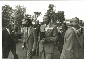 Black-and-white photograph of four men leading a march outdoors. The man in the centre is carrying a rectangular box. A group of people follows them in the background.