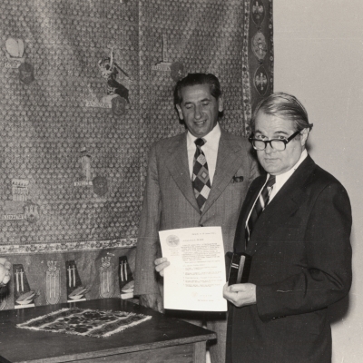 Black-and-white photograph depicting two men in suits standing around a table indoors. The man on the right is holding a document and a small urn.