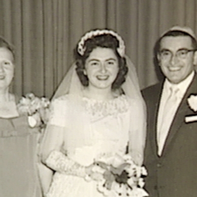 Photo en noir et blanc d'une femme debout et souriant à la caméra pendant son mariage. Elle porte une longue robe blanche, un voile et tient un bouquet de fleurs.