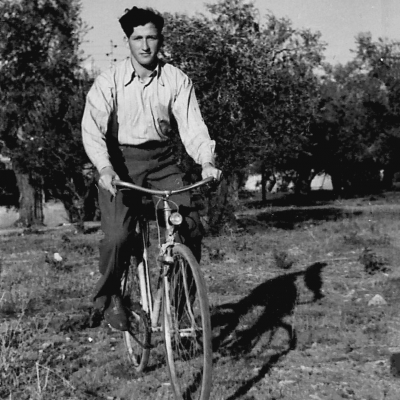 Photo en noir et blanc d'un jeune homme roulant à bicyclette à travers un champ. Il porte des pantalons foncés et une chemise à col de couleur pâle. Il y a des arbres en arrière-plan.