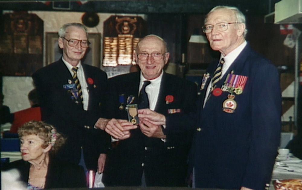 Colour photograph of three elderly men standing together and holding a military medal. The men wear suit jackets adorned in pins and medals.