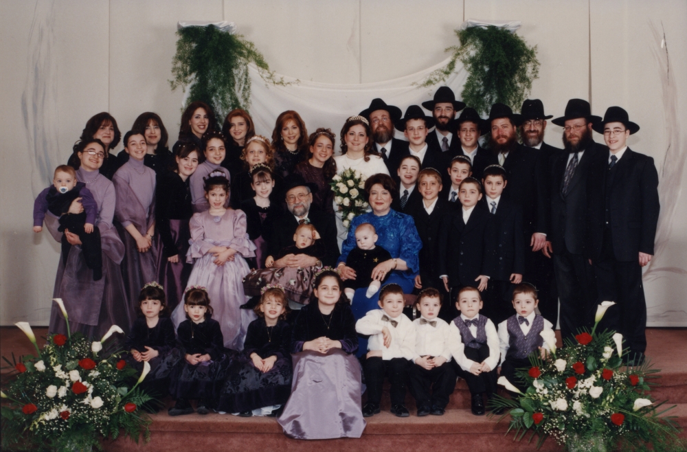 Photo en couleur d'un grand groupe d'environ 30 personnes rassemblés pour une photo de groupe de trois ou quatre rangées, certains sont assis dans la première rangée. Ils sont tous habillés de manière formelle. Les femmes portent des robes et les hommes des complets et des chapeaux. Il y a de grands bouquets de fleurs de chaque côté du groupe.