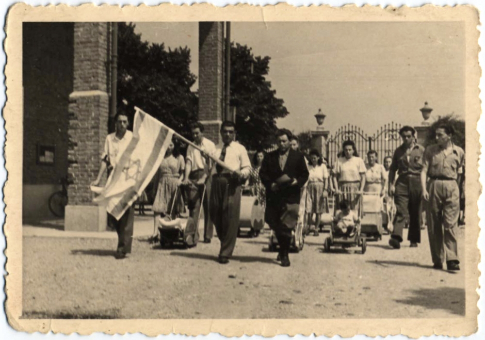 Photo en noir et blanc d'un défilé ou une marche avec un grand groupe de personnes marchant vers la caméra. Un homme à l'avant de la foule porte le drapeau d'Israël, et plusieurs femmes en arrière-plan poussent des poussettes.