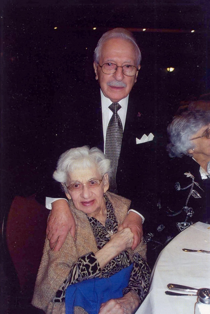 Colour photograph of two elderly people smiling at the camera. The man stands behind the woman, who is sitting at a chair, with his hands on her shoulders. She holds one of his hands.