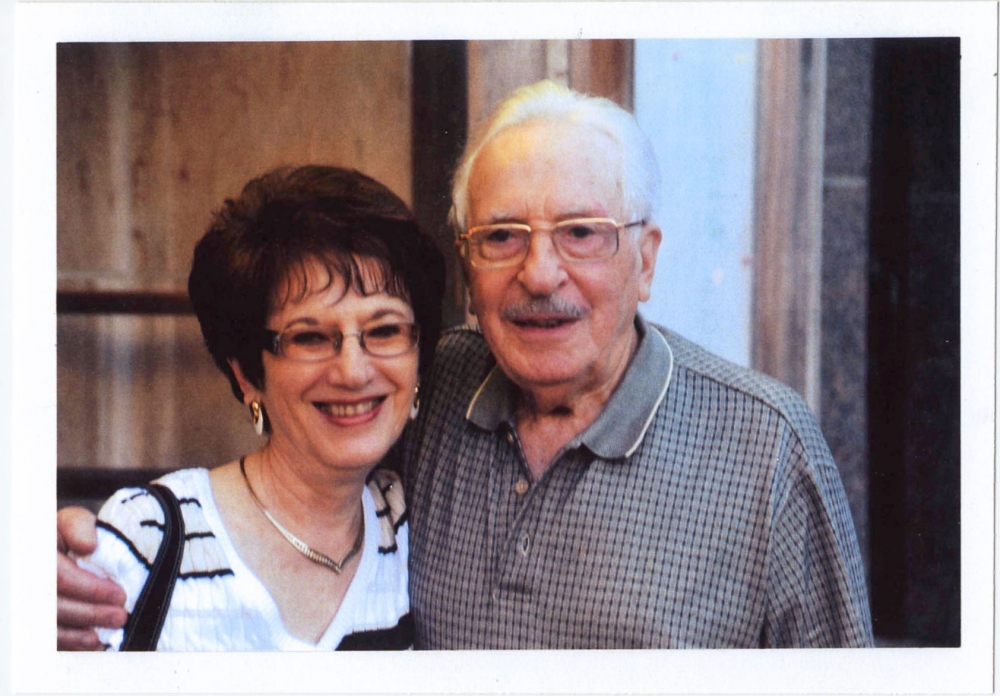 Colour photograph of an elderly couple smiling arm-in-arm at the camera. Both wear glasses, and the woman has dark brown short hair.