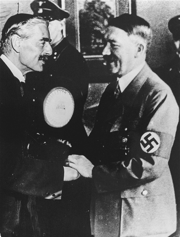 Photo en noir et blanc de deux hommes souriant et se serrant la main. Les deux hommes portent une moustache, et un des deux hommes porte un complet plus pâle avec un brassard avec une swastika.