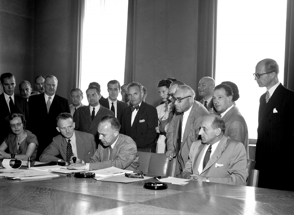 Photo en noir et blanc d'un groupe d'environ 25 personnes rassemblés ensemble derrière une grande table dans une salle de conférence. Quatre hommes sont assis à une table et un homme signe un document. Les autres se tiennent debout derrière.