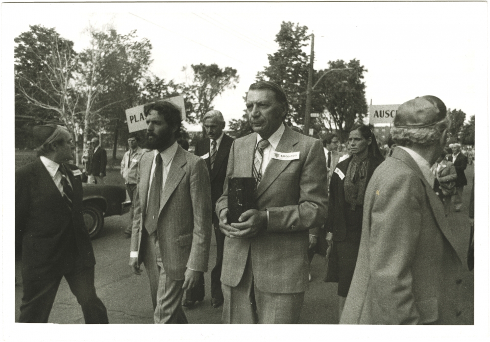 Photo en noir et blanc de quatre hommes menant une marche à l'extérieur. L'homme au centre transporte une boîte rectangulaire. Un groupe de personnes les suit à l'arrière.