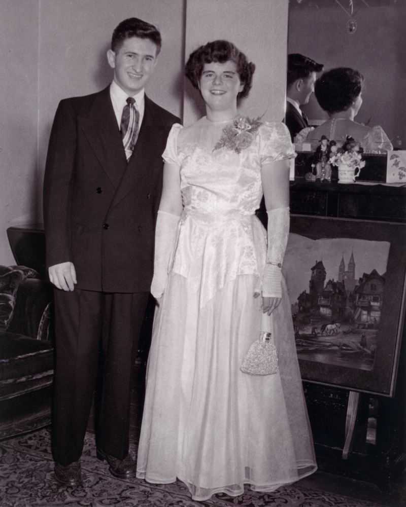 Black-and-white photograph of a young man and woman standing and smiling together indoors, in front of a mirror. The woman wears a formal long, light-coloured gown with a purse, and the man wears a suit.