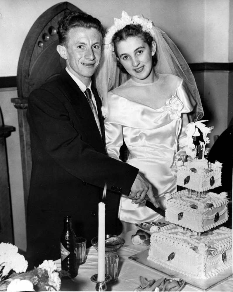 Black-and-white photograph of a man and woman standing together behind a table that holds a large, three-tiered cake. The couple, celebrating their wedding day, are cutting into the cake. They smile at the camera. The man wears a suit, and the woman wears a white silk gown with a veil.