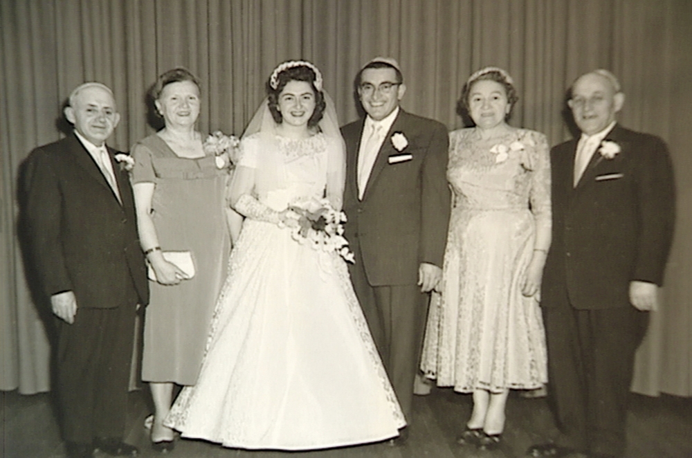 Photo en noir et blanc d'un groupe de six personnes debout en ligne et souriant à la caméra. Il semble que ce soit le mariage du couple du centre dont la femme porte une longue robe blanche, un voile et tient un bouquet de fleurs. Les trois hommes portent des complets et les deux autres femmes des robes.