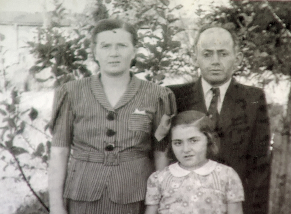 Photo en nir et blanc d'un homme et d'une femme avec leur jeune fille se tenant à l'extérieur où il y a des arbres en arrière-plan. L'homme porte un complet et la femme ainsi que la femme portent des blouses.