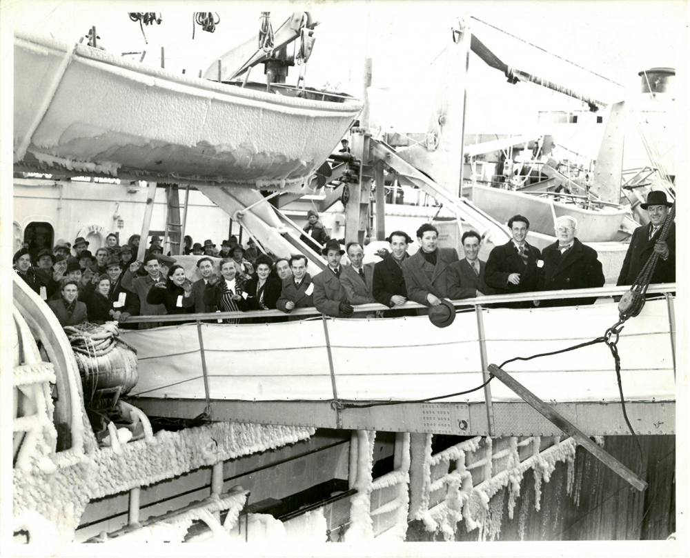 Photo en noir et blanc d'un groupe de 15 personnes, portant manteaux et chapeaux, souriant et saluant la caméra depuis la passerelle d'un navire. Plusieurs autres personnes attendent leur tour pour débarquer du navire. Un bateau de sauvetage dont les côtés sont glacés est suspendu dans le coin supérieur gauche.