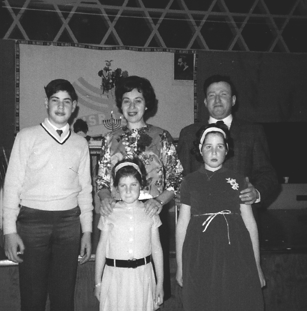 Photo en noir et blanc d'une famille de cinq, à l'intérieur, debout et souriant. La famille inclus un homme et une femme avec leur adolescent et leurs deux jeunes filles. Il y une menorah en arrière-plan.