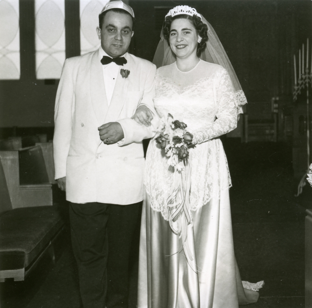 Black-and-white photograph of a man and woman, standing together arm-in-arm indoors. The couple appears to be celebrating their wedding day. The man wears a suit with a light-coloured jacket and bowtie, and the woman wears a long white gown with silk skirt, a veil, and holds a bouquet of flowers.