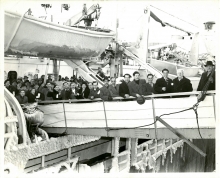 Black-and-white photograph of a group of 15 people, wearing coats and hats, smiling and waving at the camera from the gangway of a ship. Several more people wait to disembark the ship. A lifeboat is suspended in the top-left corner with ice frozen to its sides.