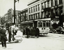 Black-and-white photograph of a street scene, taken from street level. A group of people are waiting to board a streetcar, parked on the street.