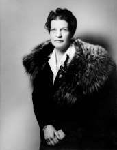 Black-and-white studio portrait photograph of a woman, pictured from the waist up, wearing a dark jacket with a fur stole. She looks directly at the camera.